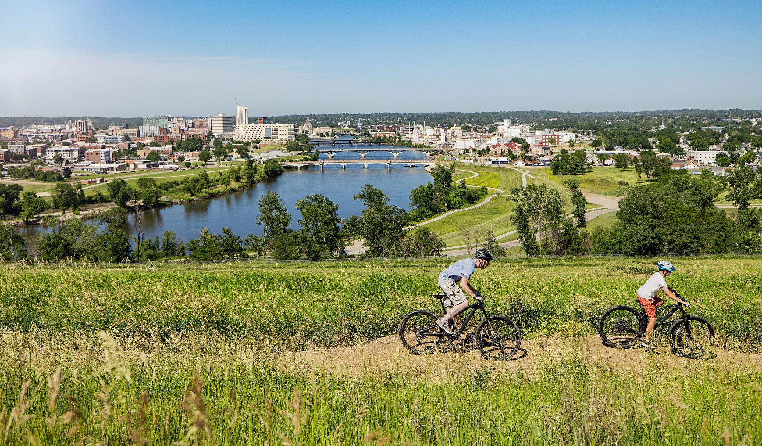 Two bikers on a trail with the Cedar Rapids skyline in the background