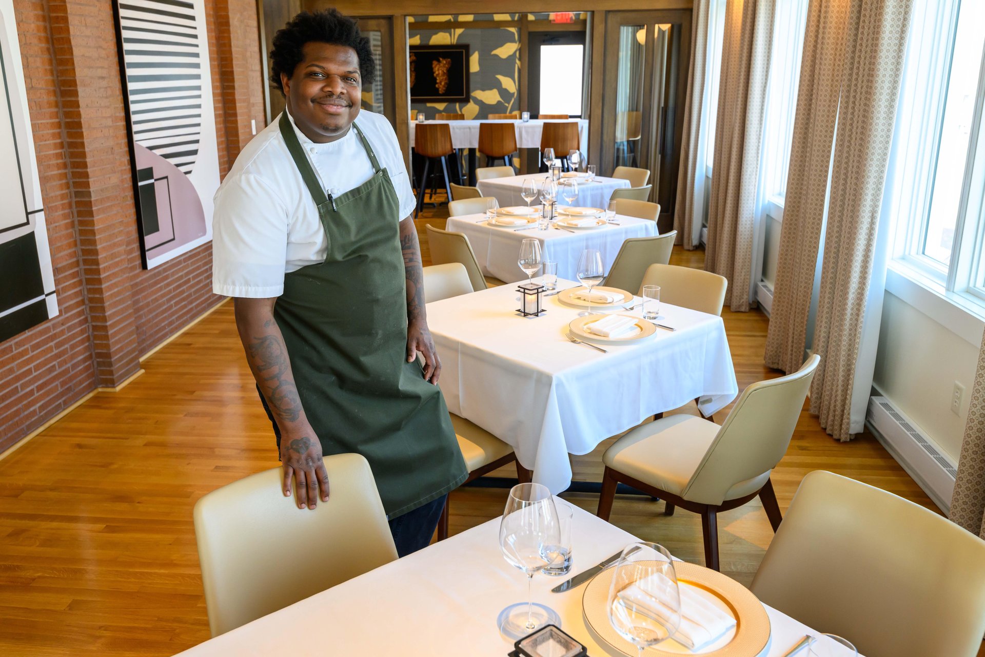 A restaurant dining area with neatly arranged tables covered in white tablecloths, set with plates, glasses, and silverware. Chef Ian Robertson is wearing a green apron and white shirt and stands near a chair, with wooden floors, large windows, and modern decor visible in the background.